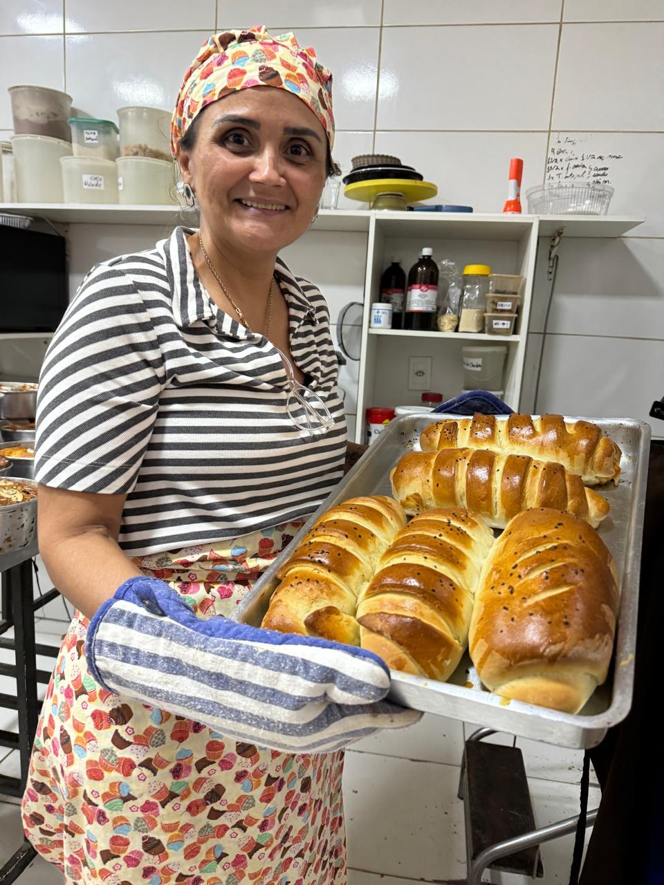Uma mulher na cozinha da Cantina do Bolo, sorrindo e segurando uma assadeira de pães